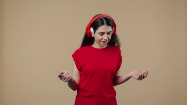 Retrato de una modelo con belleza natural disfrutando de la música en grandes auriculares rojos. Chica morena joven con el pelo largo en camiseta roja posa sobre fondo de estudio marrón. De cerca. Cámara lenta lista 59.94fps. — Vídeos de Stock