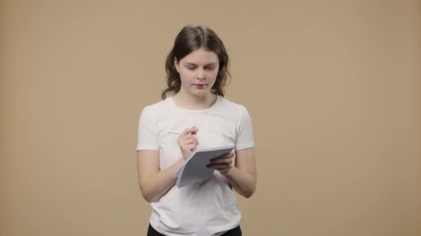 Retrato de um modelo com beleza natural é pensar com concentração e escrever ideias no caderno. Menina com cabelo encaracolado posa no fundo do estúdio marrom. Fecha. Movimento lento pronto 59.94fps. — Vídeo de Stock