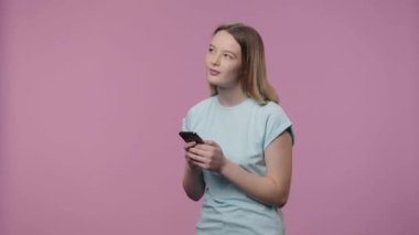 Doğal güzellikte bir modelin portresi telefonunda mesajlaşıyor. Mavi tişörtlü, uzun saçlı genç kız mor stüdyo arka planında poz veriyor. Kapatın. Yavaş çekim hazır 59.94fps.