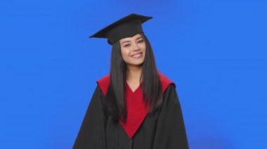 Mezuniyet kostümü giymiş kız öğrencinin portresi gülümsüyor, parmaklarıyla kalbi gösteriyor ve sonra öpüşüyor. Mavi ekranda poz veren genç esmer kadın. Kapatın. Yavaş çekim hazır 59.94fps.
