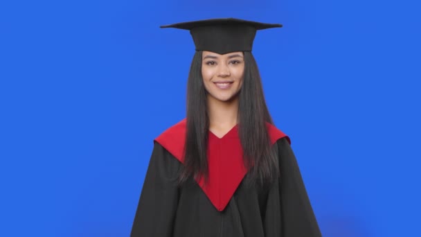 Portrait of female student in cap and gown graduation costume looking at camera and smiling. Young brunette woman posing in studio with blue screen background. Close up. Slow motion ready 59.94fps. — Stock Video