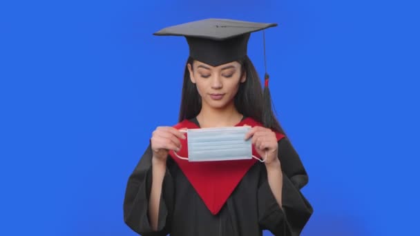 Retrato de estudante do sexo feminino em boné e vestido traje de graduação coloca máscara protetora médica. Jovem morena posando em estúdio com fundo de tela azul. Fecha. Movimento lento pronto 59.94fps. — Vídeo de Stock