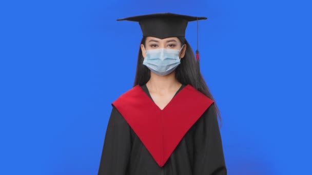 Retrato de estudante do sexo feminino em boné e vestido de graduação traje remove máscara médica protetora. Jovem morena posando em estúdio com fundo de tela azul. Fecha. Movimento lento pronto 59.94fps. — Vídeo de Stock