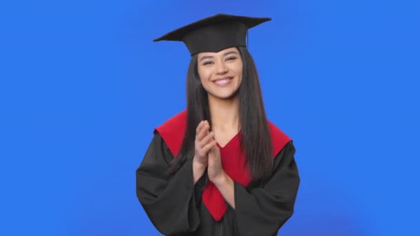 Retrato de estudante do sexo feminino em boné e vestido de graduação traje batendo palmas alegremente. Jovem morena posando em estúdio com fundo de tela azul. Fecha. Movimento lento pronto 59.94fps. — Vídeo de Stock