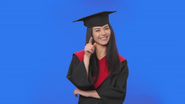 Portrait d'une étudiante en bonnet et robe graduation écoutant attentivement, hochant la tête, pointant du doigt le spectateur. Brunette posant sur fond bleu. Ferme là. Ralenti prêt 59.94fps. — Video