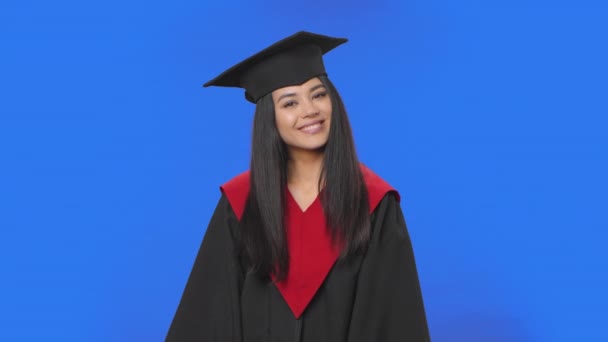 Portrait of female student in graduation costume smiling, showing heart with fingers then blowing kiss. Young brunette posing on blue screen background. Close up. Slow motion ready 59.94fps. — Stock Video