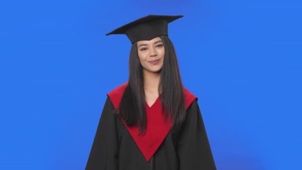 Retrato de estudante do sexo feminino em boné e vestido traje de formatura sorrindo e soprando beijo. Jovem morena posando em estúdio com fundo de tela azul. Fecha. Movimento lento pronto 59.94fps. — Vídeo de Stock