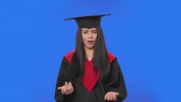 Portrait of female student in cap and gown graduation costume is indignantly talking to someone. Young brunette woman posing in studio with blue screen background. Close up. Slow motion ready 59.94fps — Stock Video