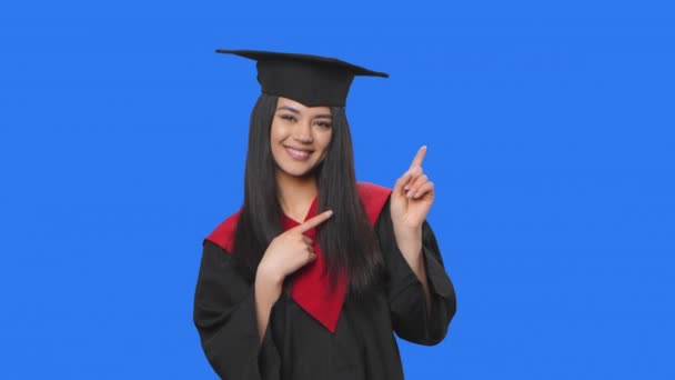 Retrato de estudante do sexo feminino no traje de graduação apontando lado mão para algo, em seguida, acena e mostra polegar como. Jovem posando no fundo da tela azul. Fecha. Movimento lento pronto 59.94fps. — Vídeo de Stock