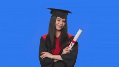 Kameraya gülümseyen ve diplomasını tutan kep ve cüppe giymiş kız öğrenci portresi. Stüdyoda poz veren genç bir kadın. Kapatın. Yavaş çekim hazır 59.94fps.