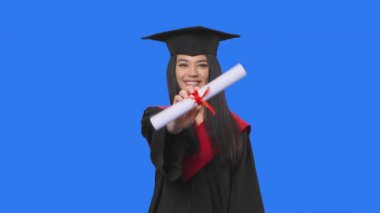 Kadın öğrenci portresi, kep ve cüppeli mezuniyet kostümü, elinde diploma ve baş parmak gösterme. Stüdyoda poz veren genç bir kadın. Kapatın. Yavaş çekim hazır 59.94fps.