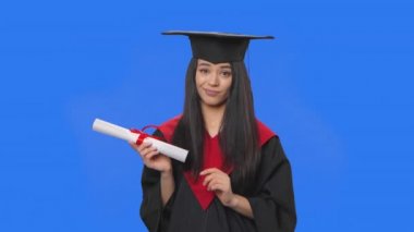 Kadın öğrenci portresi, kep ve cübbe içinde mezuniyet kostümü, diploma elinde ve baş parmağını aşağı gösteriyor. Stüdyoda poz veren genç bir kadın. Kapatın. Yavaş çekim hazır 59.94fps.