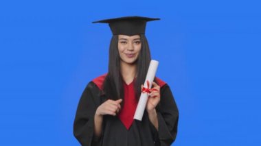 Kadın öğrenci portresi, kep ve cübbe içinde mezuniyet kostümü, elinde diploma ve iyi niyet gösterisi. Stüdyoda poz veren genç bir kadın. Kapatın. Yavaş çekim hazır 59.94fps.