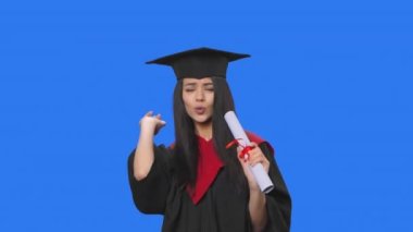 Mezuniyet kostümü giymiş, elinde diplomayla dans edip neşeyle şarkı söyleyen bir kız öğrenci portresi. Stüdyoda poz veren genç bir kadın. Kapatın. Yavaş çekim hazır 59.94fps.