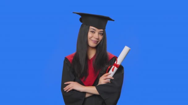Retrato de una estudiante con traje de graduación de gorra y vestido sonriendo a la cámara y sosteniendo su diploma. Mujer joven posando en el estudio con fondo de pantalla azul. De cerca. Cámara lenta lista 59.94fps. — Vídeos de Stock