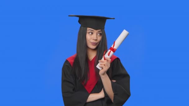 Retrato de estudante do sexo feminino no traje de graduação olhando atenciosamente para o seu diploma, então se alegra há uma ideia. Jovem posando no fundo da tela azul. Fecha. Movimento lento pronto 59.94fps — Vídeo de Stock