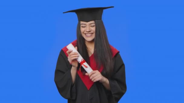 Retrato de estudante do sexo feminino em boné e vestido traje de formatura, segurando seu diploma e é muito feliz. Jovem posando em estúdio com fundo de tela azul. Fecha. Movimento lento pronto 59.94fps. — Vídeo de Stock