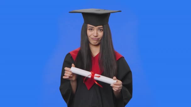 Retrato de estudante do sexo feminino em boné e vestido traje de formatura, segurando seu diploma e está muito chateado. Jovem posando em estúdio com fundo de tela azul. Fecha. Movimento lento pronto 59.94fps. — Vídeo de Stock