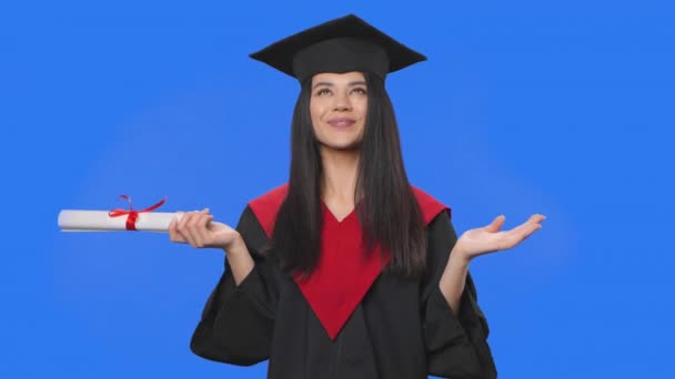 Retrato de estudante do sexo feminino em boné e vestido traje de graduação, segurando seu diploma, suspira com alívio e sorrisos. Jovem posando no fundo da tela azul. Fecha. Movimento lento pronto 59.94fps. — Vídeo de Stock