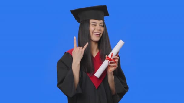 Portrait of female student in cap and gown graduation costume, holding diploma and making rock gesture. Young woman posing in studio with blue screen background. Close up. Slow motion ready 59.94fps. — Stock Video