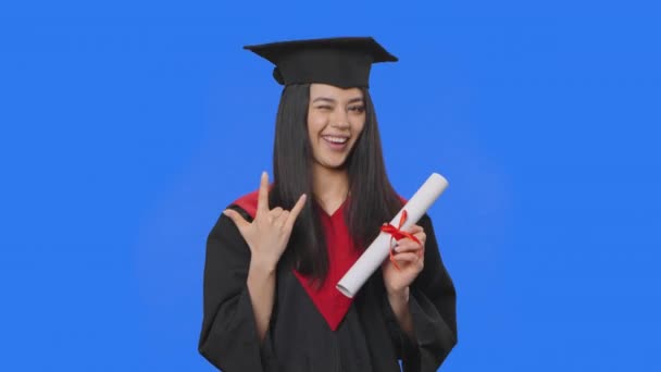 Portrait d'une étudiante en costume de fin d'études en casquette et en robe, titulaire d'un diplôme et faisant un geste rock. Jeune femme posant en studio avec fond bleu. Ferme là. Ralenti prêt 59.94fps. — Video