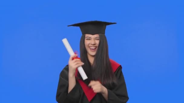 Portrait d'une étudiante en costume de fin d'études en casquette et en robe, titulaire d'un diplôme et dansant joyeusement. Jeune femme posant en studio avec fond bleu. Ferme là. Ralenti prêt 59.94fps. — Video