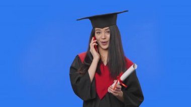 Mezuniyet kostümü giymiş bir kız öğrencinin portresi, elinde diploma, elinde akıllı bir telefonla konuşuyor. Stüdyoda mavi ekran arka planda poz veren esmer bir kadın. Kapatın. Yavaş çekim hazır 59.94fps.