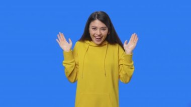 Kameraya hayranlık ve hayranlıkla bakan güzel bir esmerin portresi. Stüdyoda mavi ekran arka planı olan sarı kazaklı genç bir kadın. Kapatın. Yavaş çekim hazır 59.94fps.