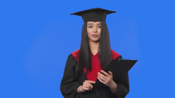 Retrato de una estudiante en traje de graduación, contando información y sosteniendo una carpeta negra en las manos. Mujer joven posando en el estudio con fondo de pantalla azul. De cerca. Cámara lenta lista 59.94fps. — Vídeo de stock