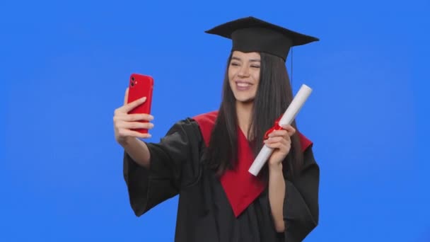 Portret van een vrouwelijke studente in afstudeerkostuum, met een diploma en een videogesprek via de telefoon. Jonge vrouw poseren in de studio met blauwe achtergrond scherm. Sluit maar af. Slow motion klaar 59,94fps. — Stockvideo