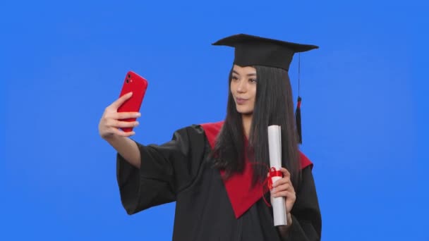 Retrato de estudante do sexo feminino em traje de graduação, segurando diploma e fazendo selfie no telefone móvel. Jovem posando em estúdio com fundo de tela azul. Fecha. Movimento lento pronto 59.94fps. — Vídeo de Stock