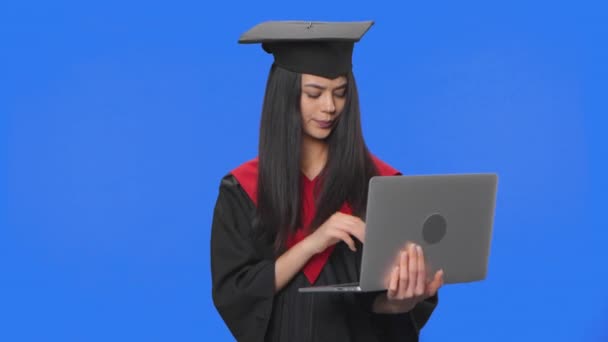 Retrato de estudiante femenina en traje de graduación de la gorra y el vestido, se comunica en línea utilizando un ordenador portátil. Mujer joven posando en el estudio con fondo de pantalla azul. De cerca. Cámara lenta lista 59.94fps. — Vídeos de Stock