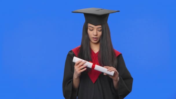 Портрет студентки-жінки в шапці і сукні випускного костюму, що тримає диплом, а потім викидає його. Молода жінка позує на фоні синього екрану в студії. Близько. Повільний рух готовий 59.94fps — стокове відео