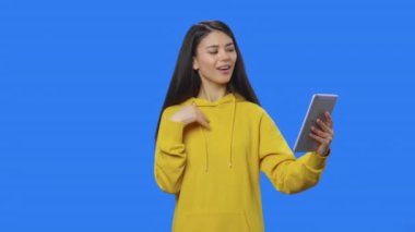 Tablet kullanarak görüntülü konuşma yapan güzel esmerin portresi. Sarı kazaklı, uzun saçlı, stüdyoda poz veren, mavi ekran arka planlı genç bir kadın. Kapatın. Yavaş çekim hazır 59.94fps.