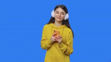 Büyük beyaz kulaklıklı, telefonla müzik dinleyen güzel esmerin portresi. Stüdyoda mavi ekran arka planı olan sarı kazaklı genç bir kadın. Kapatın. Yavaş çekim hazır 59.94fps.