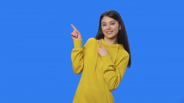 Porträtt av vacker brunett pekar sida hand för något och visar tummen som. ung kvinna i gul tröja poserar i studio med blå skärm bakgrund. Närbild. Slow motion klar 59,94fps. — Stockvideo