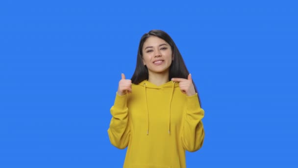 Portrét krásné brunetky ukazující prstem nahoru a ukazující palec. Mladá žena s dlouhými vlasy ve žluté mikině pózuje ve studiu s modrým pozadí obrazovky. Zavřít. Zpomalení připraveno 59.94fps — Stock video