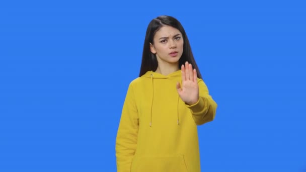 Portret brunetki ściśle gestykulującej kształtem dłoni, co oznacza zaprzeczenie "NIE". Młoda kobieta w żółtej bluzie, pozująca w studio z niebieskim tłem. Zamknij drzwi. Gotowy do zwolnień 59.94fps. — Wideo stockowe
