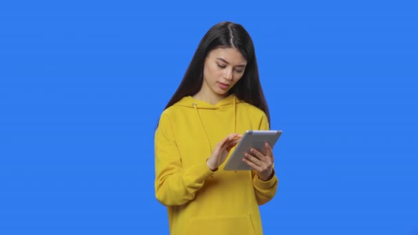 Πορτρέτο των όμορφων μελαχρινή προβολή πληροφοριών σε ένα ψηφιακό tablet. Νεαρή γυναίκα με μακριά μαλλιά σε κίτρινο φούτερ ποζάρουν στο στούντιο με φόντο μπλε οθόνη. Κλείσε. Αργή κίνηση έτοιμη 59.94fps. — Αρχείο Βίντεο