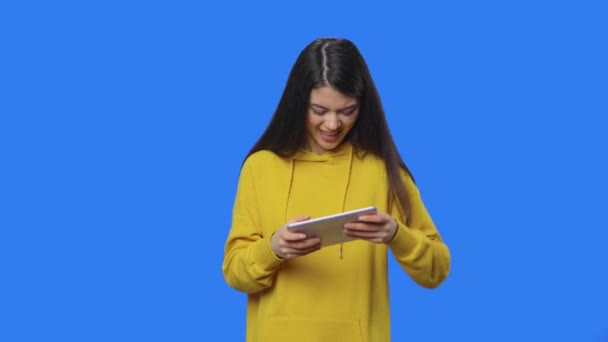 Portrét krásné brunetky nadšeně hraje hru na digitálním tabletu. Mladá žena ve žluté mikině pózuje ve studiu s modrým pozadí obrazovky. Zavřít. Zpomalení připraveno 59.94fps. — Stock video