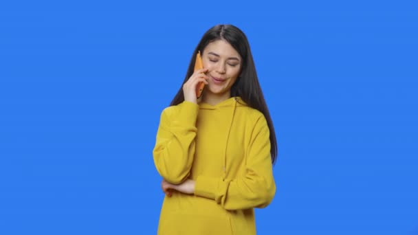 Portret van een mooie brunette die praat op een smartphone. Jonge vrouw met lang haar in gele sweater poserend in studio met blauwe achtergrond. Sluit maar af. Slow motion klaar 59,94fps. — Stockvideo