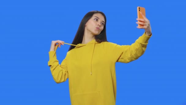 Retrato de la bonita morena haciendo selfie en el teléfono móvil y luego mirando fotos. Mujer joven en sudadera amarilla posando en estudio con fondo de pantalla azul. De cerca. Cámara lenta lista 59.94fps. — Vídeo de stock