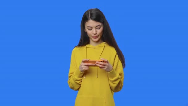 Portrét krásné brunetky nadšeně hraje hru na smartphonu. Mladá žena ve žluté mikině pózuje ve studiu s modrým pozadí obrazovky. Zavřít. Zpomalení připraveno 59.94fps. — Stock video