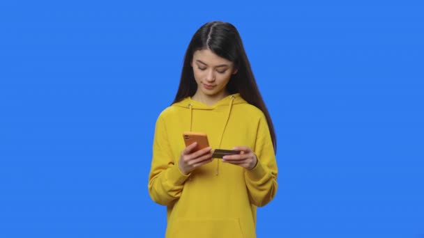漂亮黑发的画像是用信用卡和电话在网上购买的。身穿黄色运动衫的年轻女子摆出一副蓝屏背景的画室姿势.靠近点慢动作准备59.94fps. — 图库视频影像