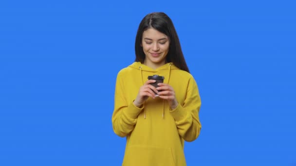 Portræt af smuk brunette nyder kaffe. Ung kvinde med langt hår i gul sweatshirt poserende i studie med blå skærm baggrund. Tæt på. Langsom bevægelse klar 59.94fps. – Stock-video