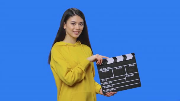 Portret van een mooie brunette met klapbord en glimlach. Jonge vrouw met lang haar in gele sweater poserend in studio met blauwe achtergrond. Sluit maar af. Slow motion klaar 59,94fps. — Stockvideo