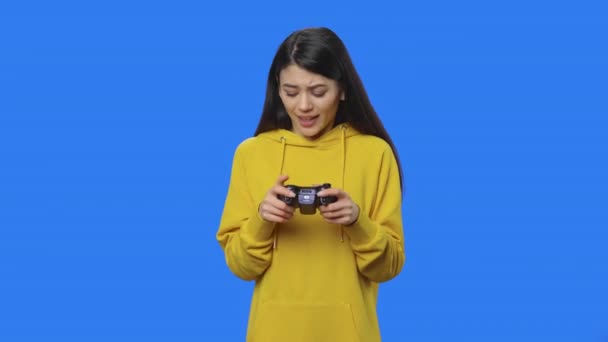 Retrato de morena jogando videogame usando controle sem fio e se alegrando na vitória. Jovem mulher de camisola amarela posando no fundo da tela azul. Fecha. Movimento lento pronto 59.94fps. — Vídeo de Stock