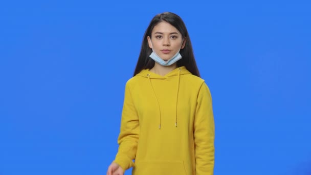 Portret van een mooie brunette die laat zien hoe je een medisch masker moet dragen. Jonge vrouw in geel sweatshirt poseren in studio met blauwe achtergrond. Sluit maar af. Slow motion klaar 59,94fps. — Stockvideo