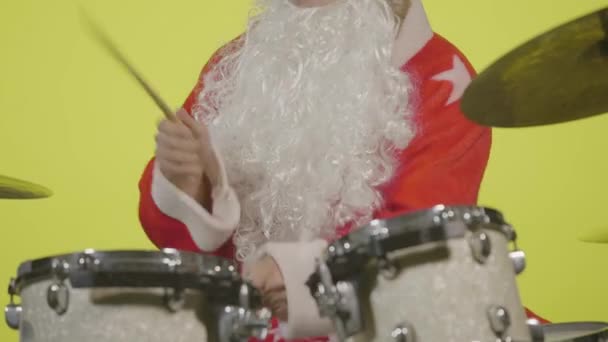 Święty Mikołaj w świątecznym stroju z brodą i okularami przeciwsłonecznymi gra na perkusji na żółtym tle studio. Człowiek w stroju karnawałowym grzywka bębny i blachy perkusyjne do tworzenia muzyki. Zamknij drzwi. Zwolniony ruch. — Wideo stockowe