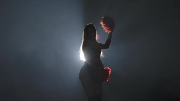 Young female cheerleader with orange pompoms in uniform is dancing on black smoky studio background with backlight. Silhouette of dancer performs cheering dance. Close up. Slow motion ready 59.94fps. — Stock Video